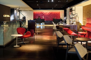 A modern lobby with a reception desk, red chairs, a piano, and a staircase.