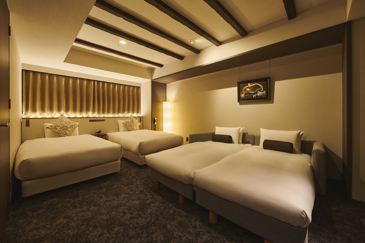 A hotel room with two single beds, a sofa bed, a large window with curtains, a wall-mounted lamp, and a framed picture.