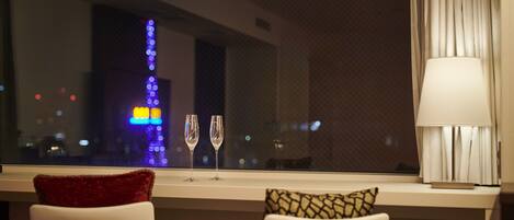 Habitación de hotel con vistas a las luces de la ciudad a través de una ventana, dos sillones con cojines y una mesa con lámpara.