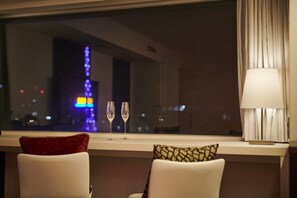A hotel room with a view of city lights through a window, two chairs with pillows, and a table with a lamp.