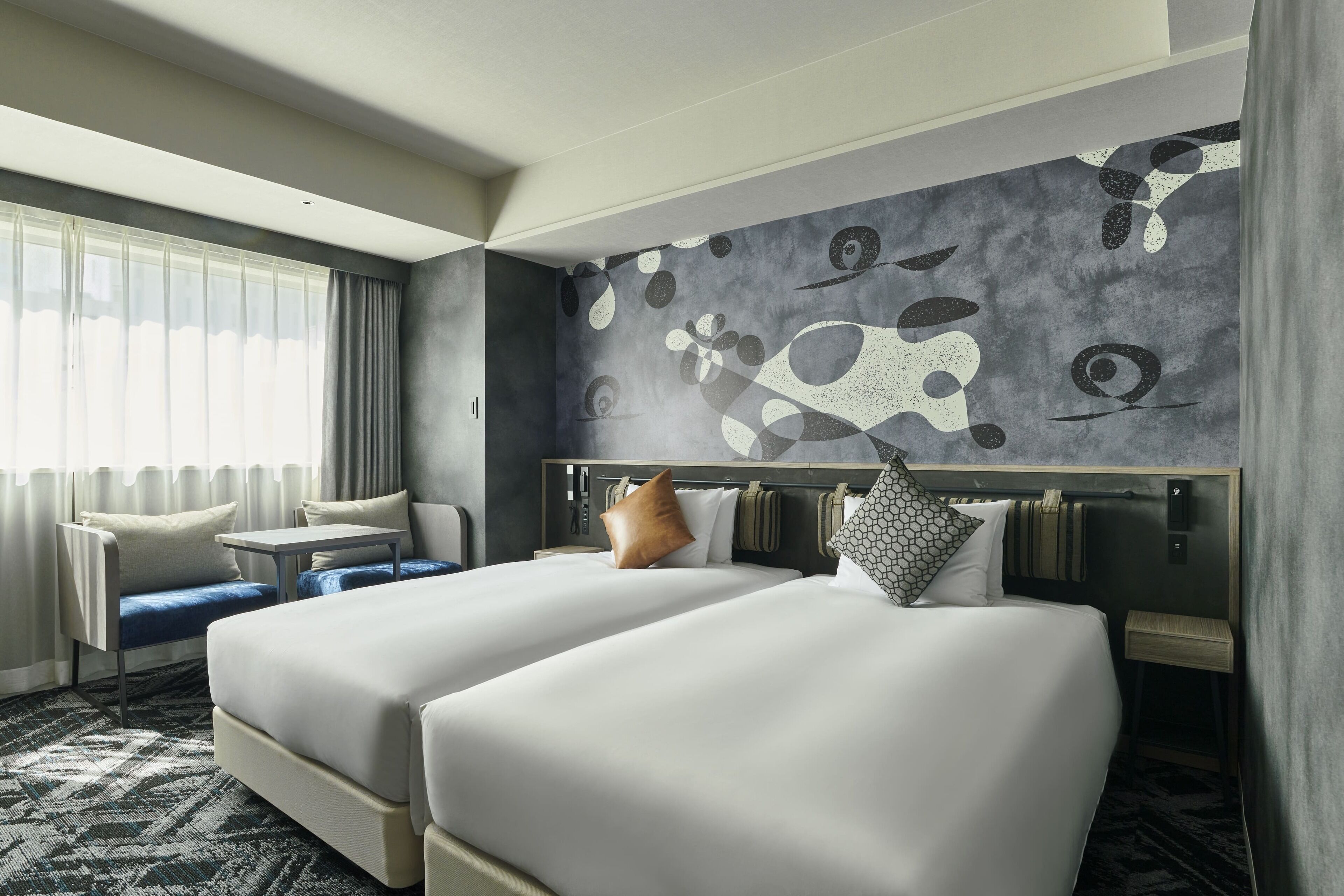 Habitación de hotel con dos camas, un sofá, una mesita pequeña y un mural abstracto grande en la pared.