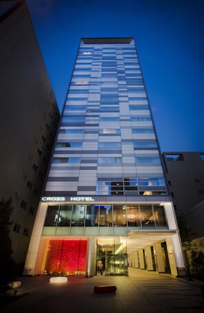 A modern hotel with a glass facade, labeled "CROSS HOTEL," featuring a prominent entrance with a red accent wall and a well-lit interior.