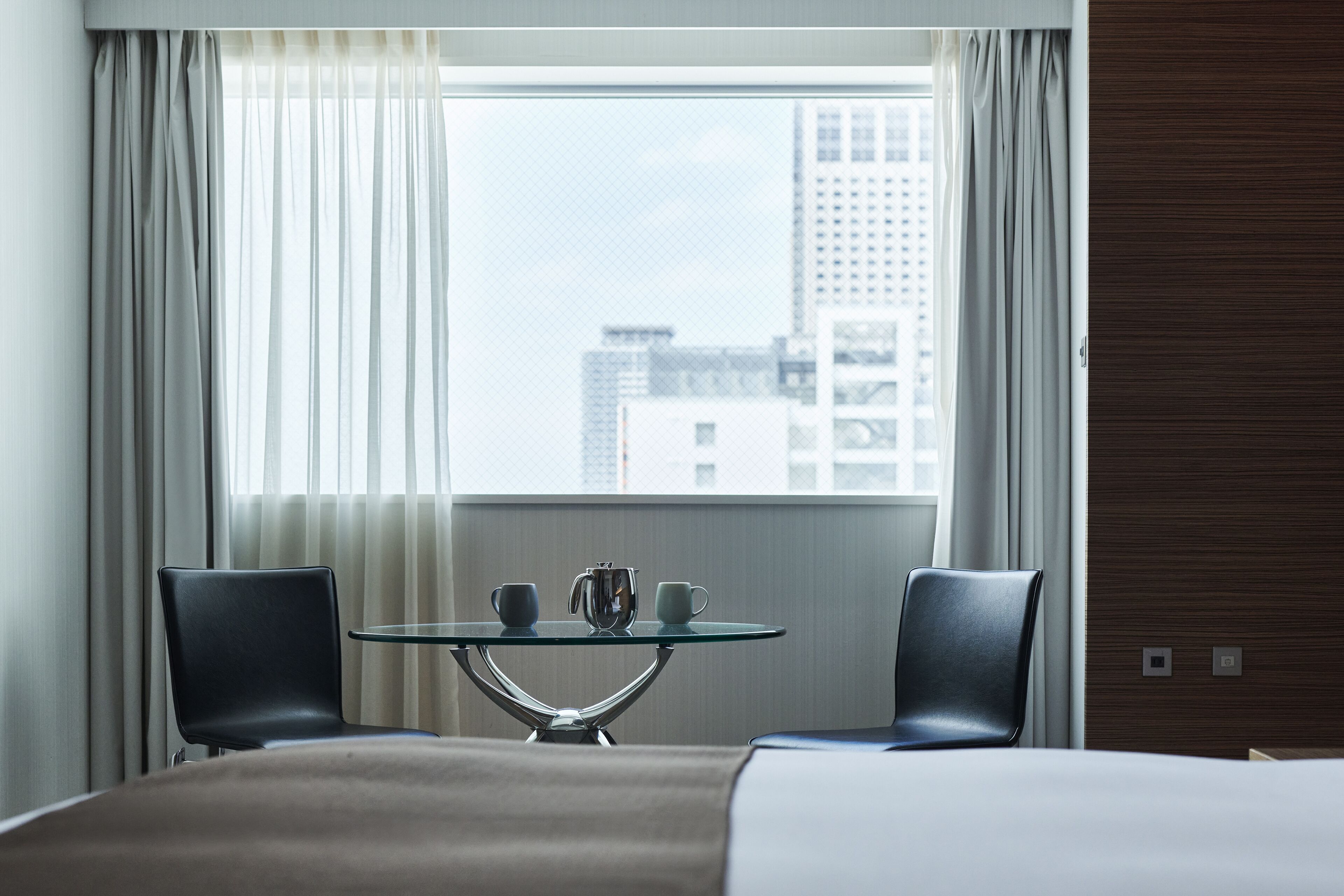 Ein Hotelzimmer mit einem Bett, zwei Stühlen, einem Glastisch mit Teekanne und Tassen sowie einem Fenster mit Vorhängen.