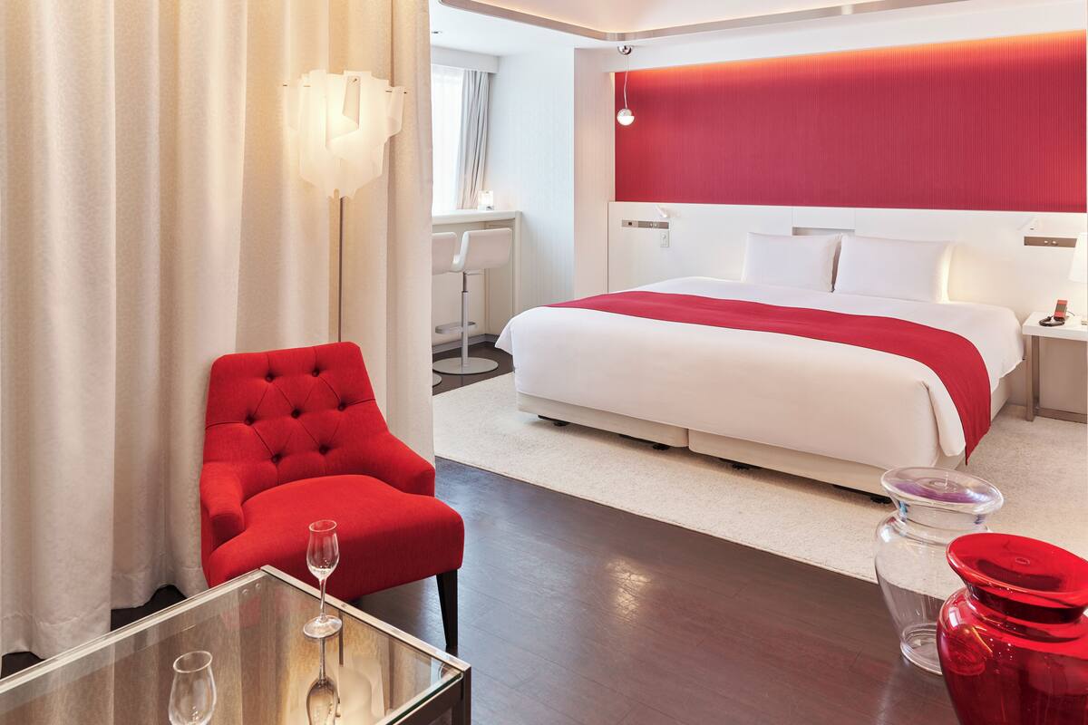 A hotel room with a large bed, a red armchair, a glass table with two wine glasses, and a red vase.