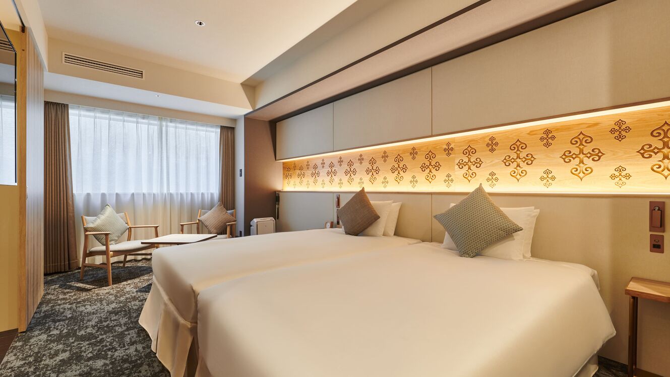 A hotel room with two beds, a sitting area with a chair, a desk, and a large window with curtains.