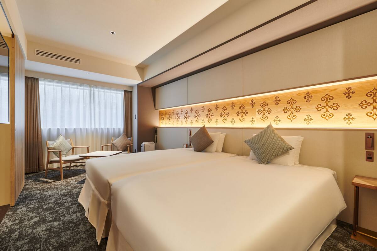 A hotel room with two beds, a desk, a chair, and a large window with curtains.