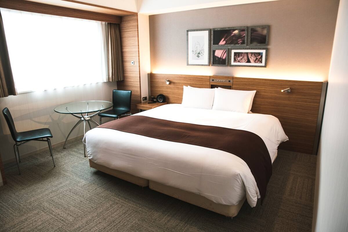 A hotel room with a large bed, a small round table, a chair, and a window with curtains.