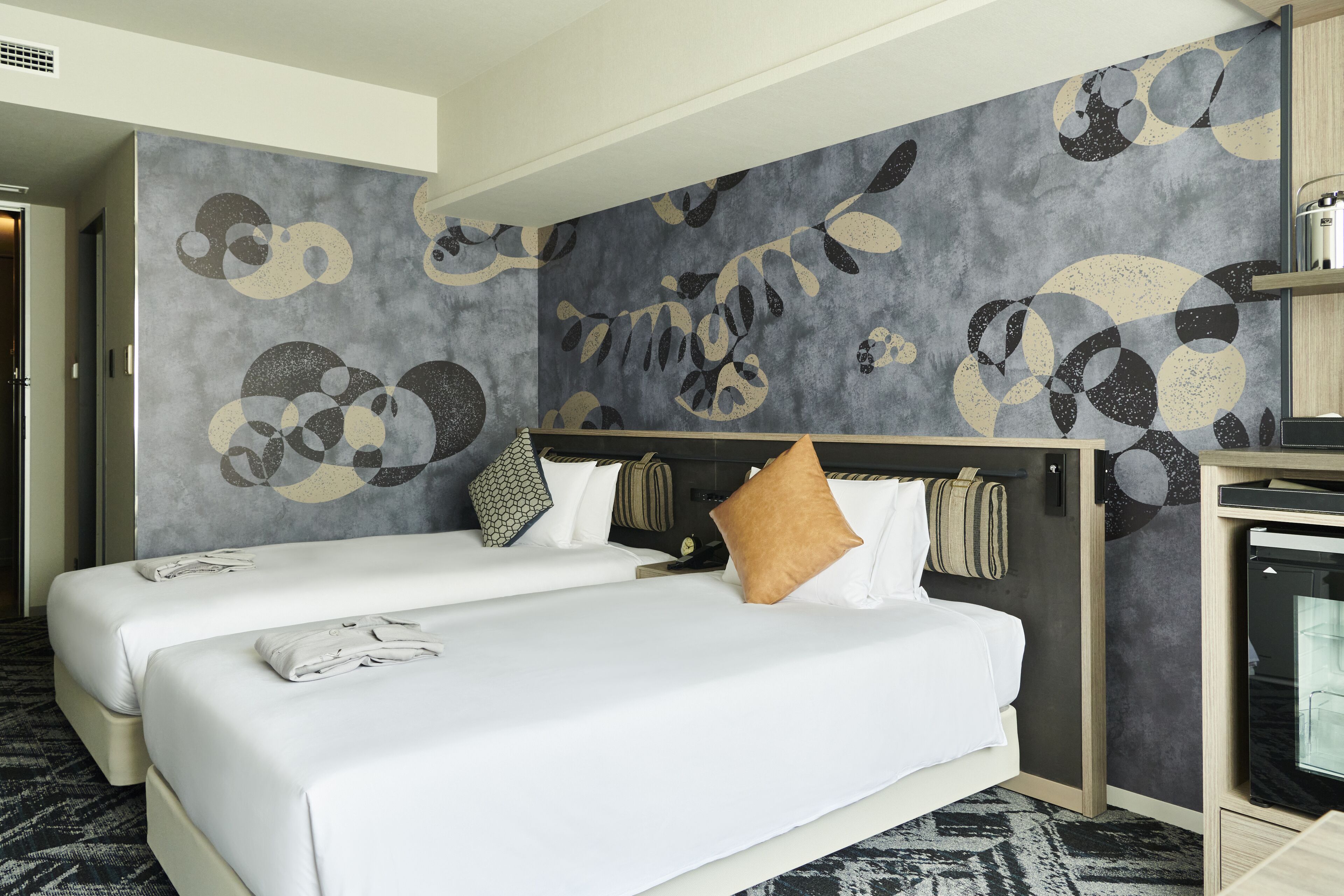 A hotel room with two beds, a large abstract wall mural, a small kitchenette, and a wardrobe.