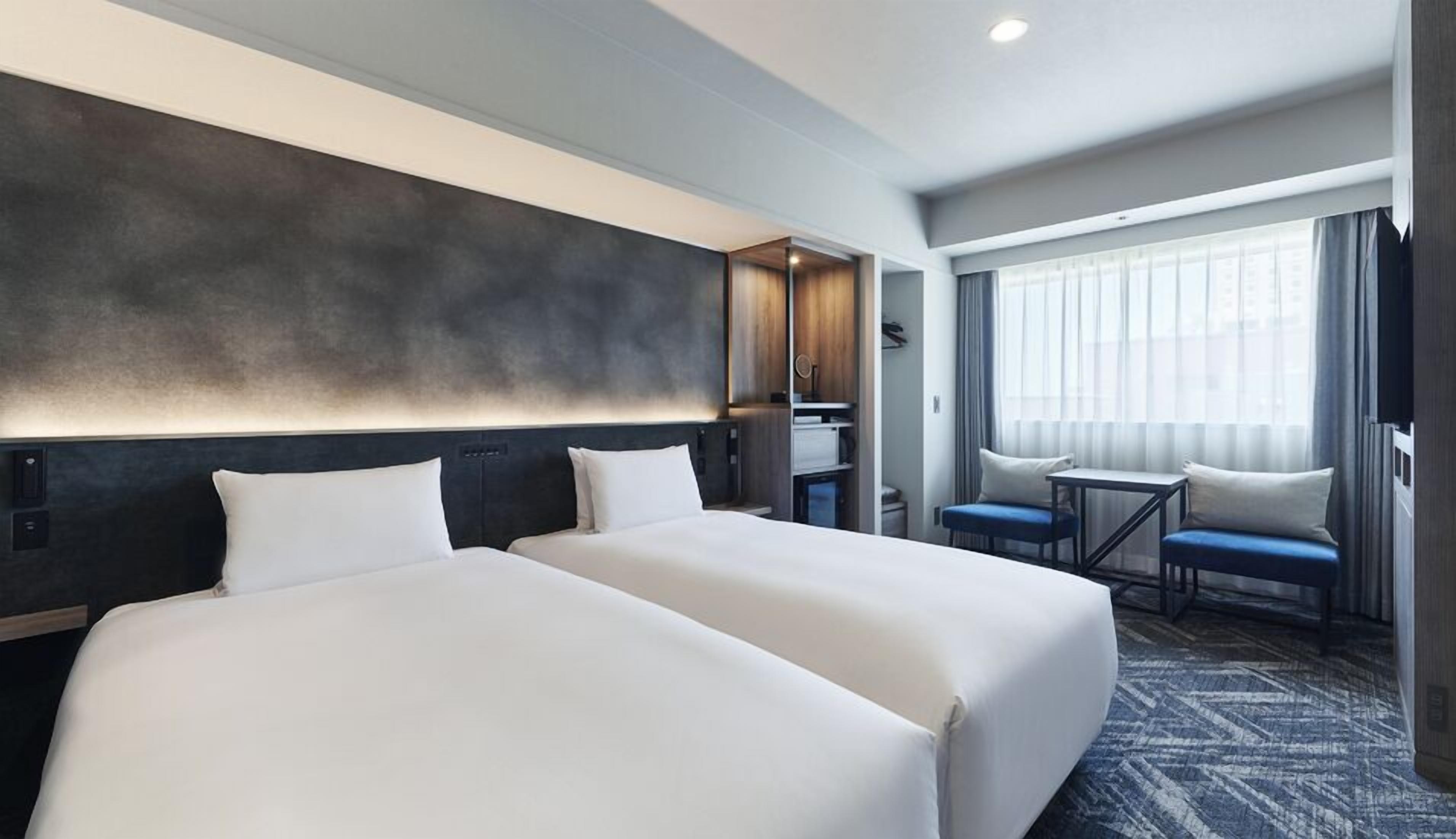 A hotel room with two beds, a desk, a chair, and a large window with curtains.