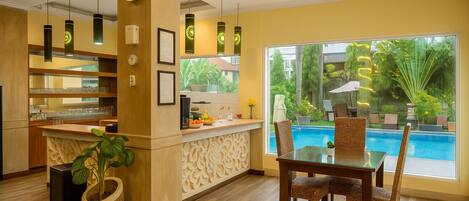 A modern hotel lobby with a pool view, a reception desk, and a dining area with wicker chairs.