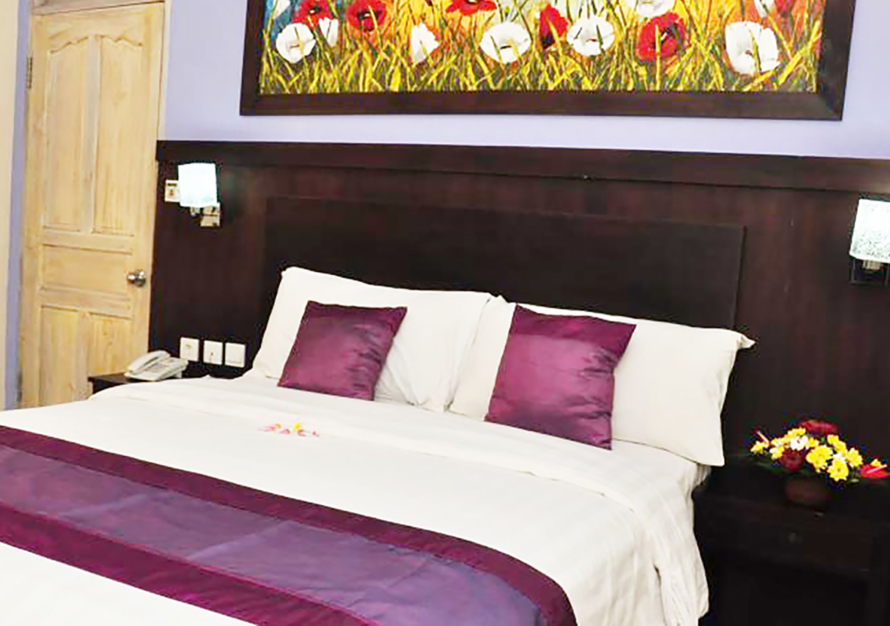 A hotel room with a large bed, a wooden headboard, a nightstand with a phone, and a painting of flowers on the wall.