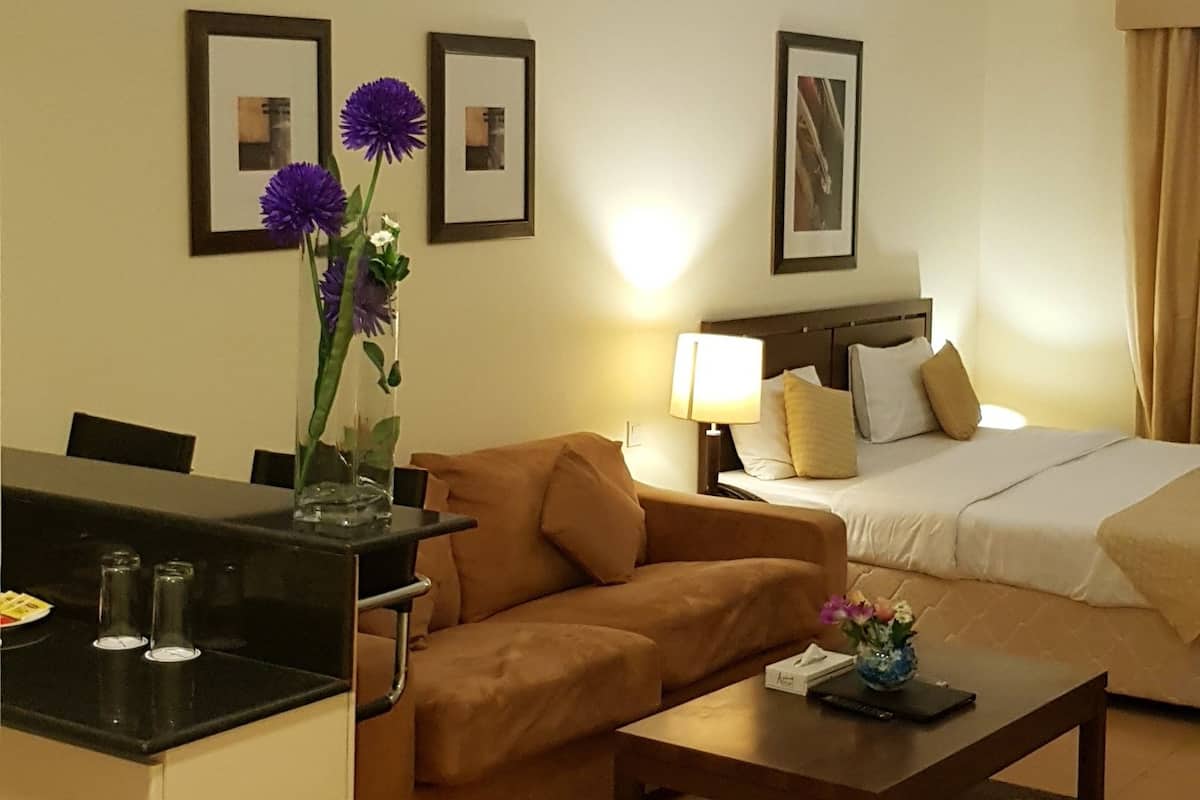 A hotel room with a sofa, a small table, a bed, and a desk with a chair. There are framed pictures on the wall and a trash can near the door.