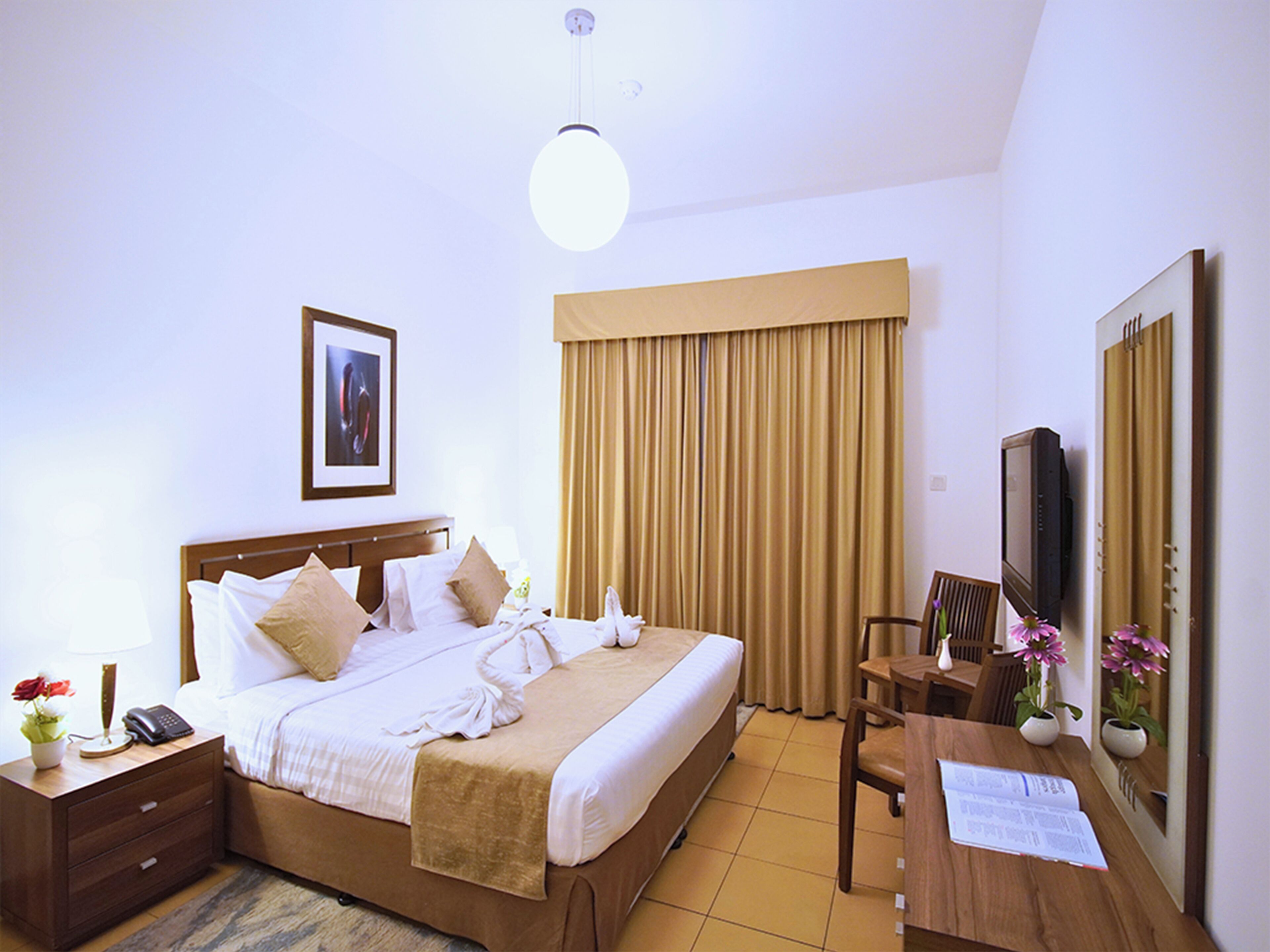 A hotel room with a bed, bedside table, lamp, TV, and a desk with a chair.