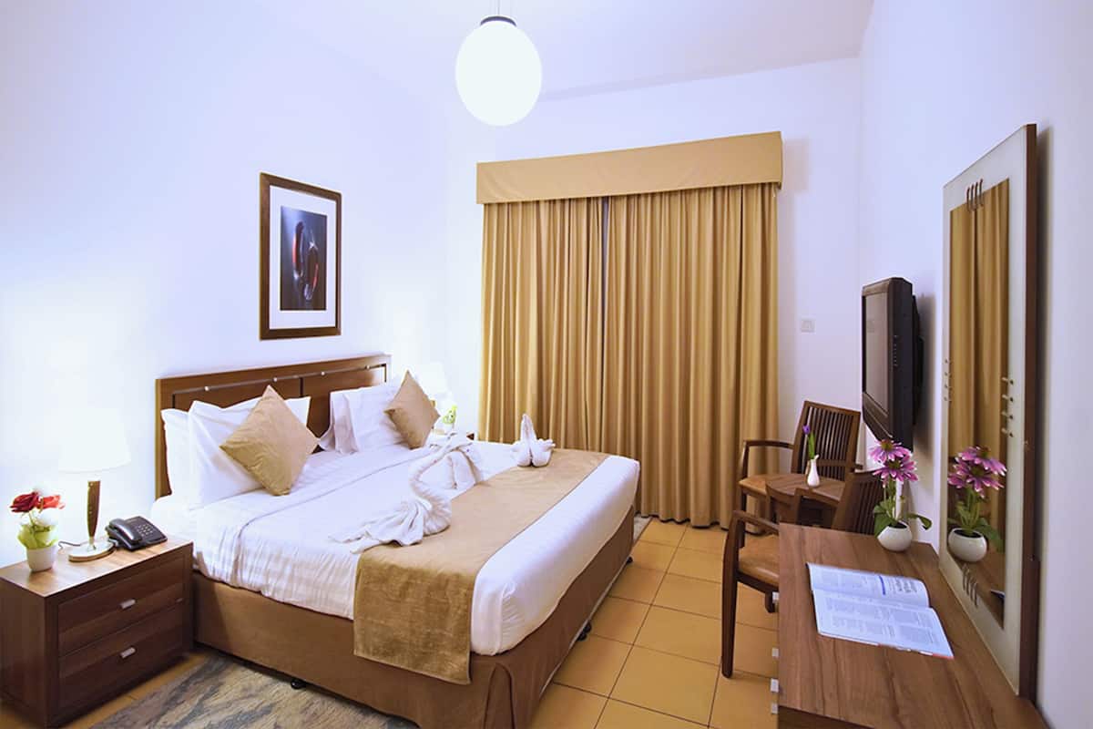 A hotel room with a bed, bedside table, lamp, TV, and a desk with a chair.