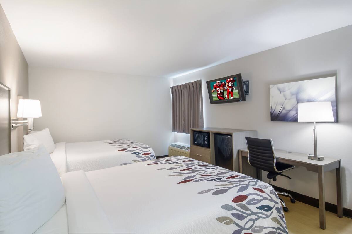 A hotel room with two beds, a desk, a chair, a TV, and a lamp.