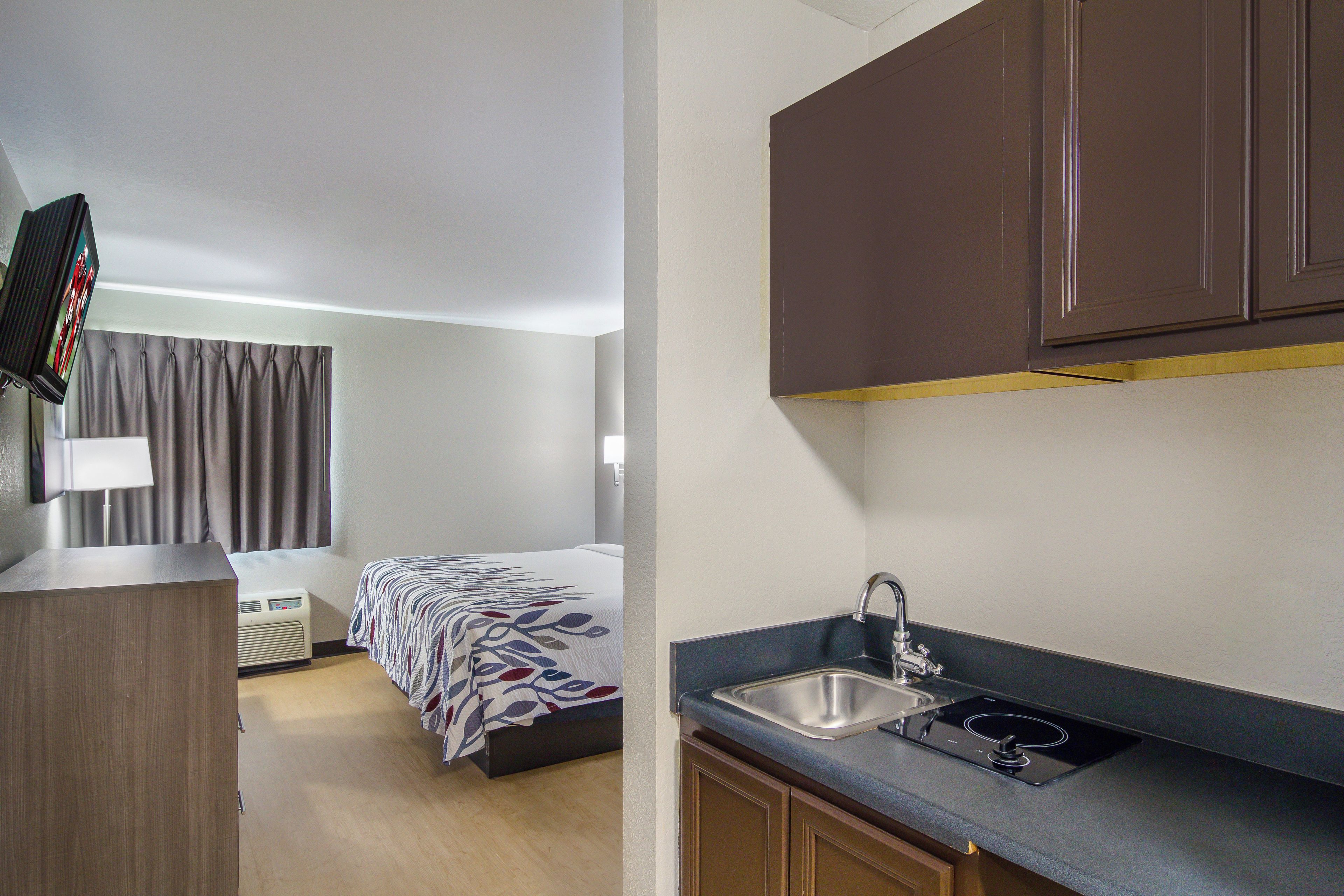Una habitación de hotel compacta con kitchenette, una cama con colcha estampada, televisor y ventana con cortinas.