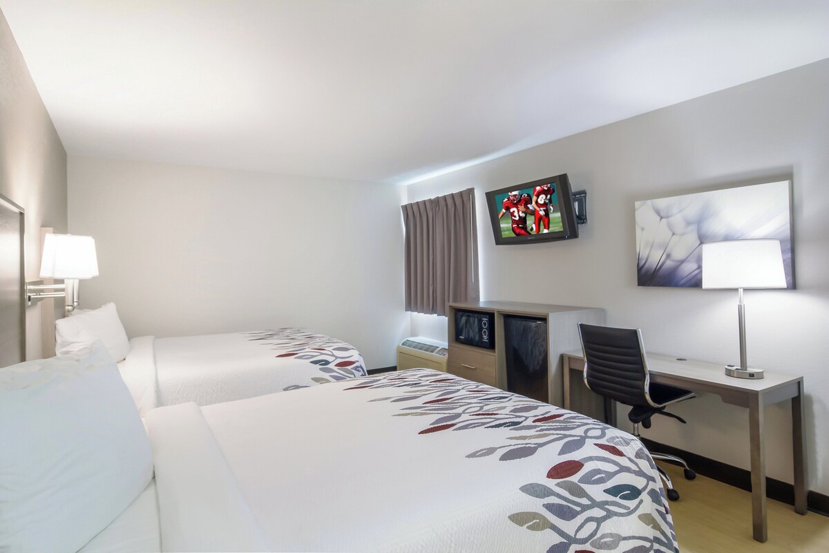 A hotel room with two beds, a desk, a chair, a TV, and a lamp.