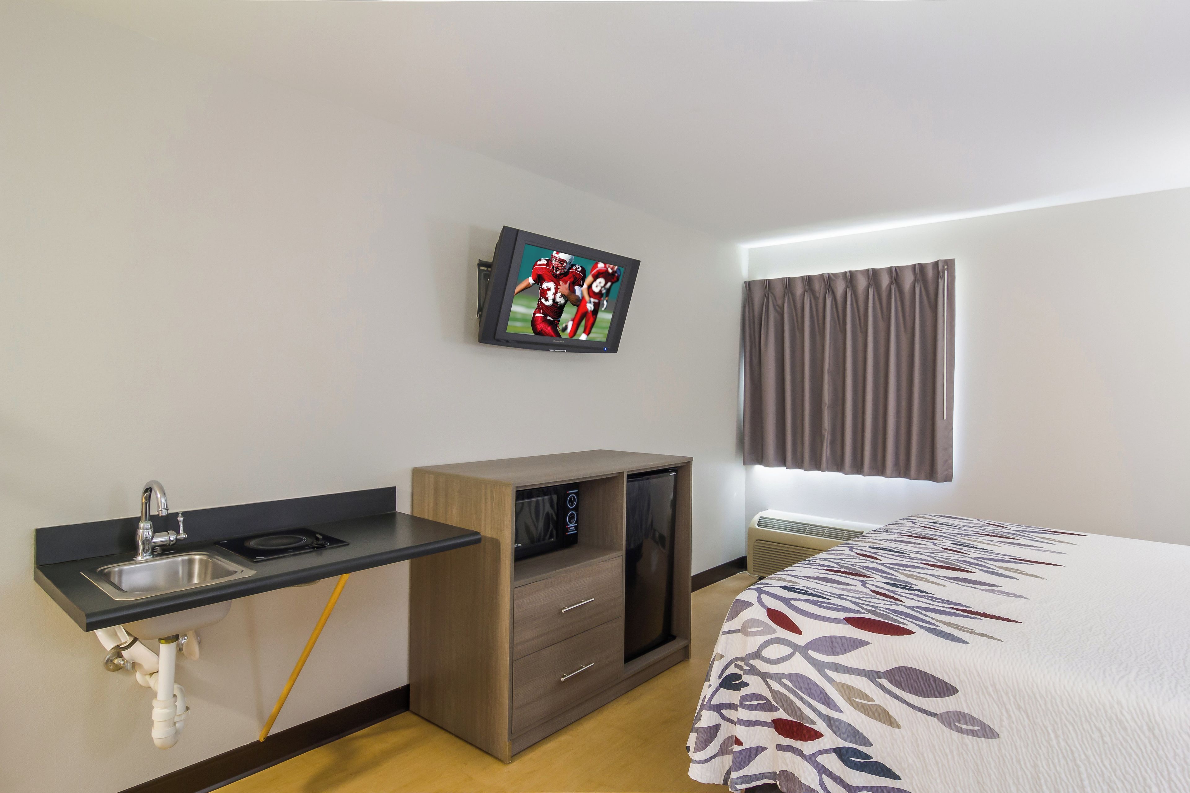A hotel room with a bed, a TV mounted on the wall, a small kitchenette, and a window with curtains.