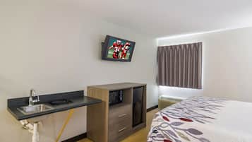 Habitación de hotel con cama, televisión montada en la pared, kitchenette pequeña y ventana con cortinas.