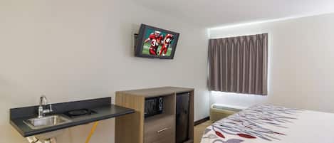 Una camera d'albergo con un letto, una TV a parete, un angolo cottura e una finestra con tende.