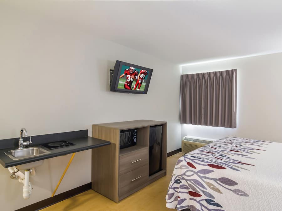 Habitación de hotel con cama, televisión montada en la pared, kitchenette pequeña y ventana con cortinas.