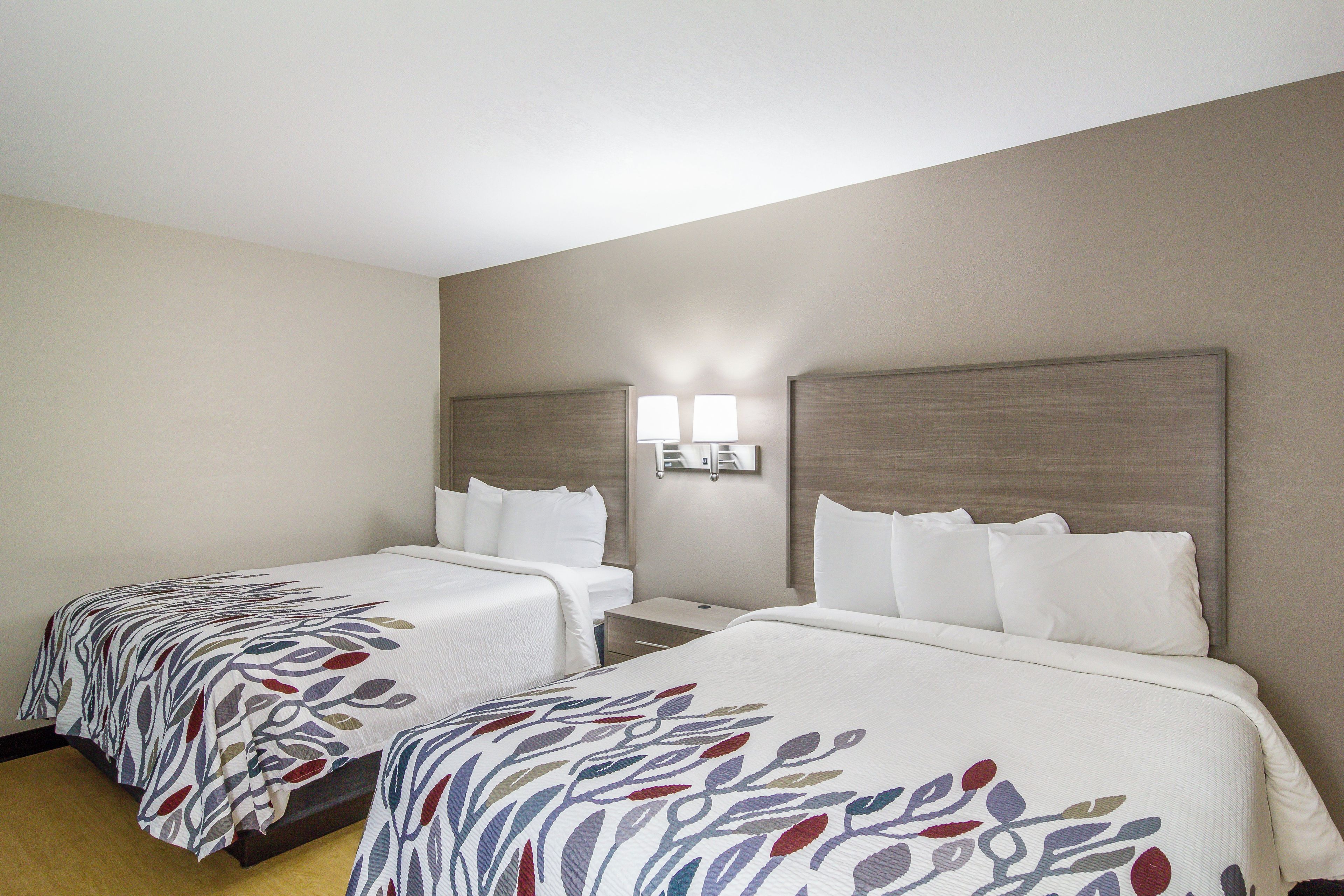 A hotel room with two beds, each with a patterned bedspread, a nightstand, and a wall-mounted lamp between the beds.