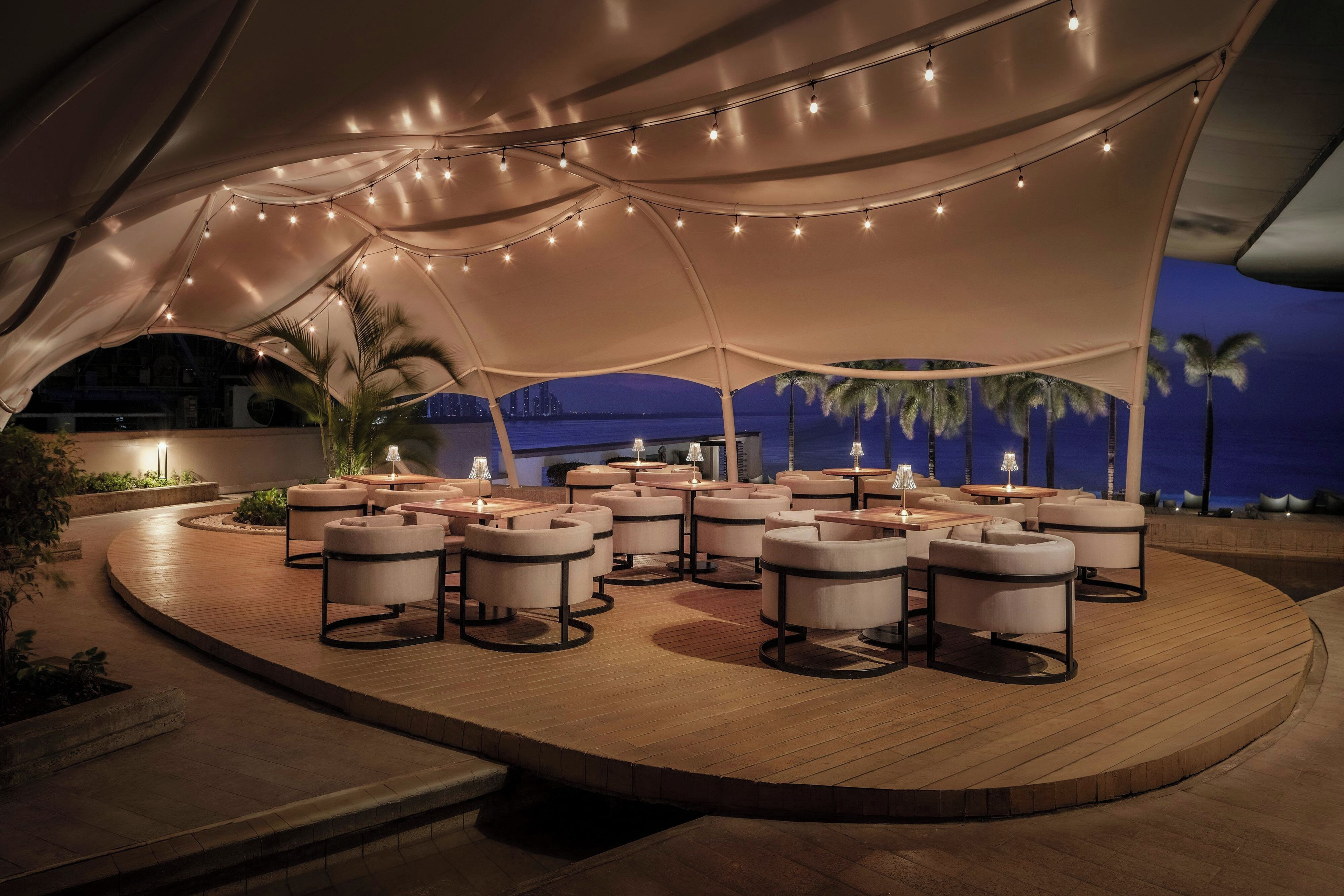 An outdoor seating area with round tables and chairs, under a large tent with string lights.