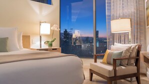 Een moderne hotelkamer met een groot bed, een stoel, een nachtkastje met lamp en uitzicht op de stad in de schemering.