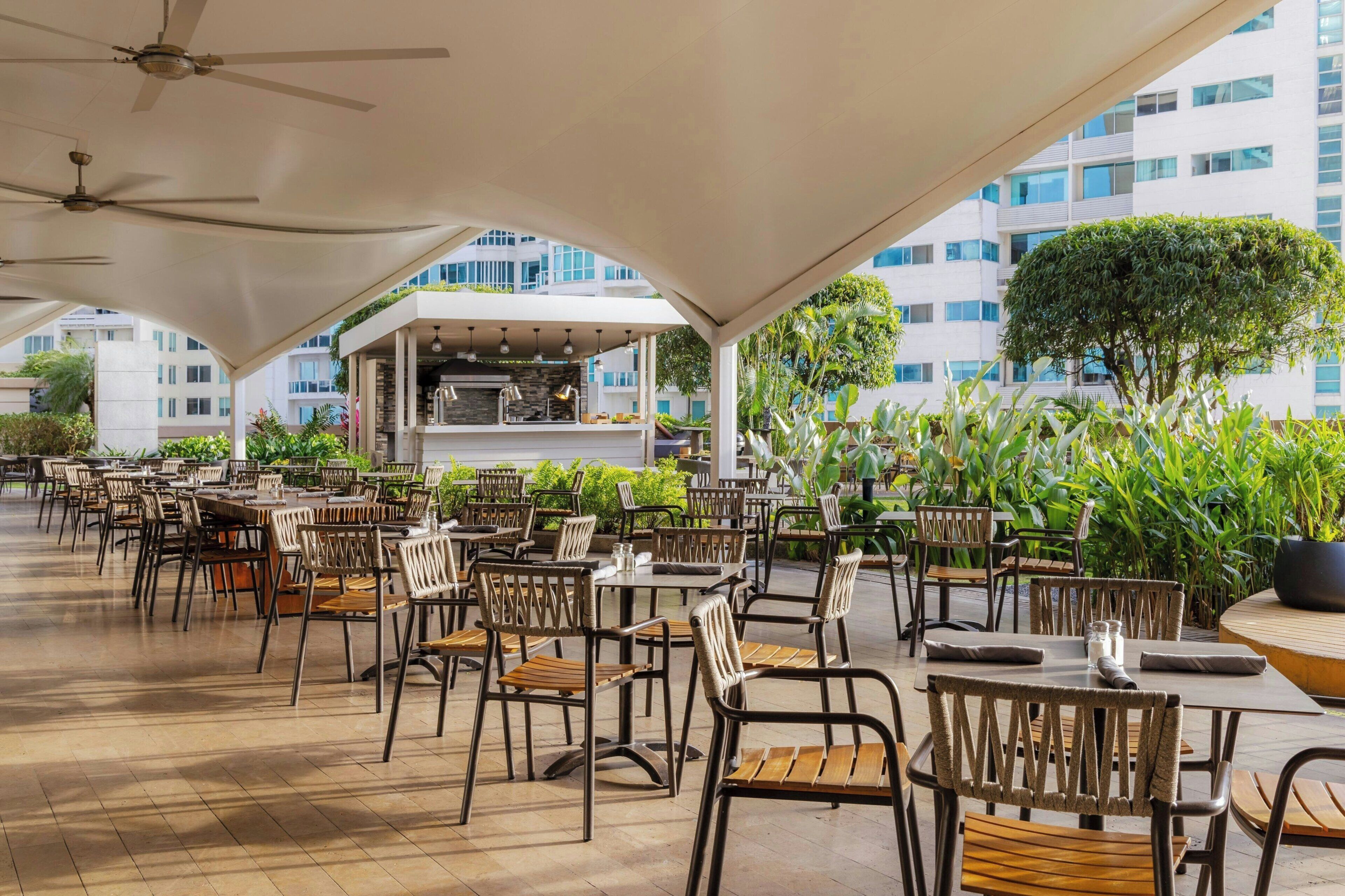 Un área de comedor al aire libre con mesas y sillas, una barra central y vista a un edificio moderno.
