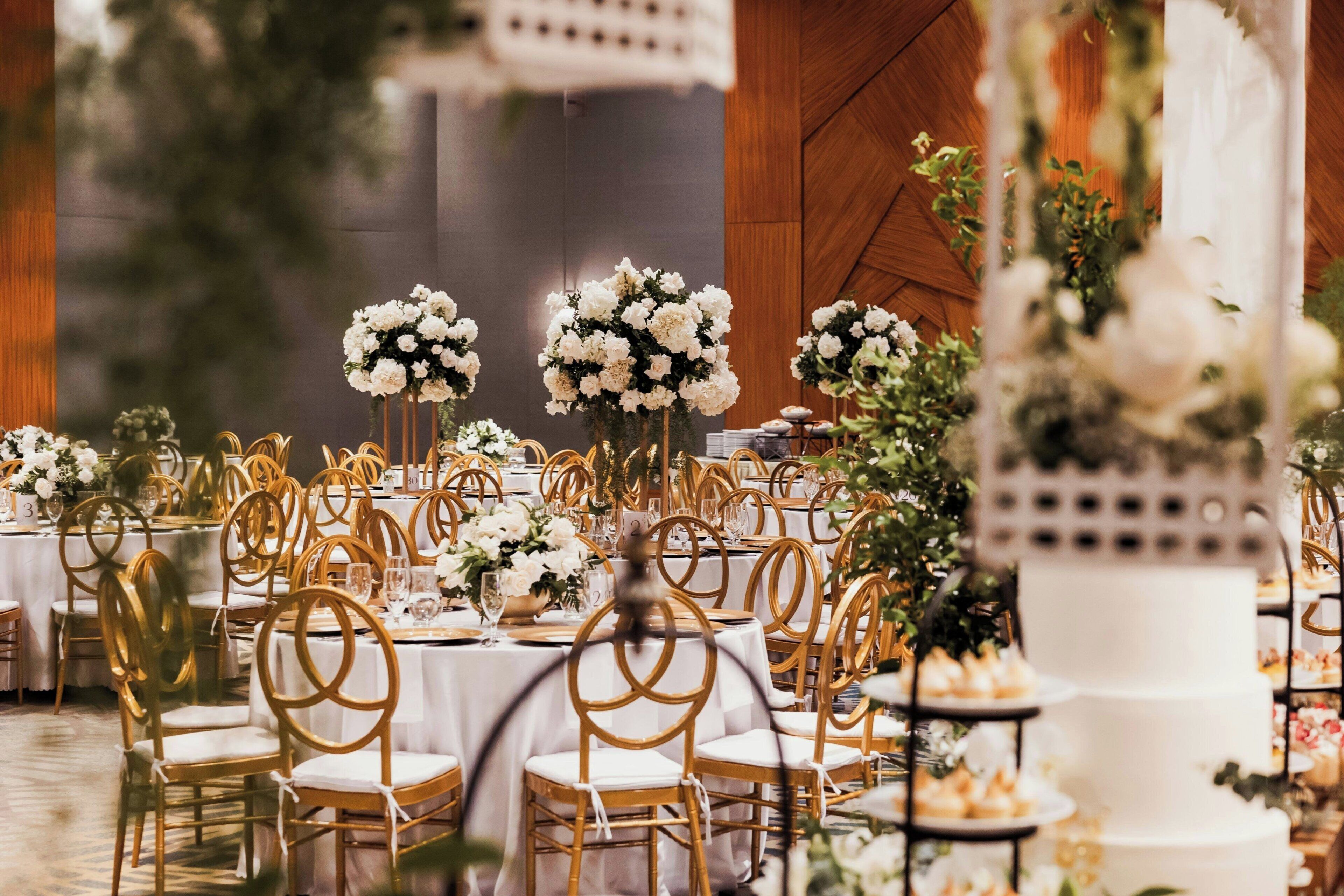 A banquet hall with round tables, gold chairs, and white floral centerpieces.