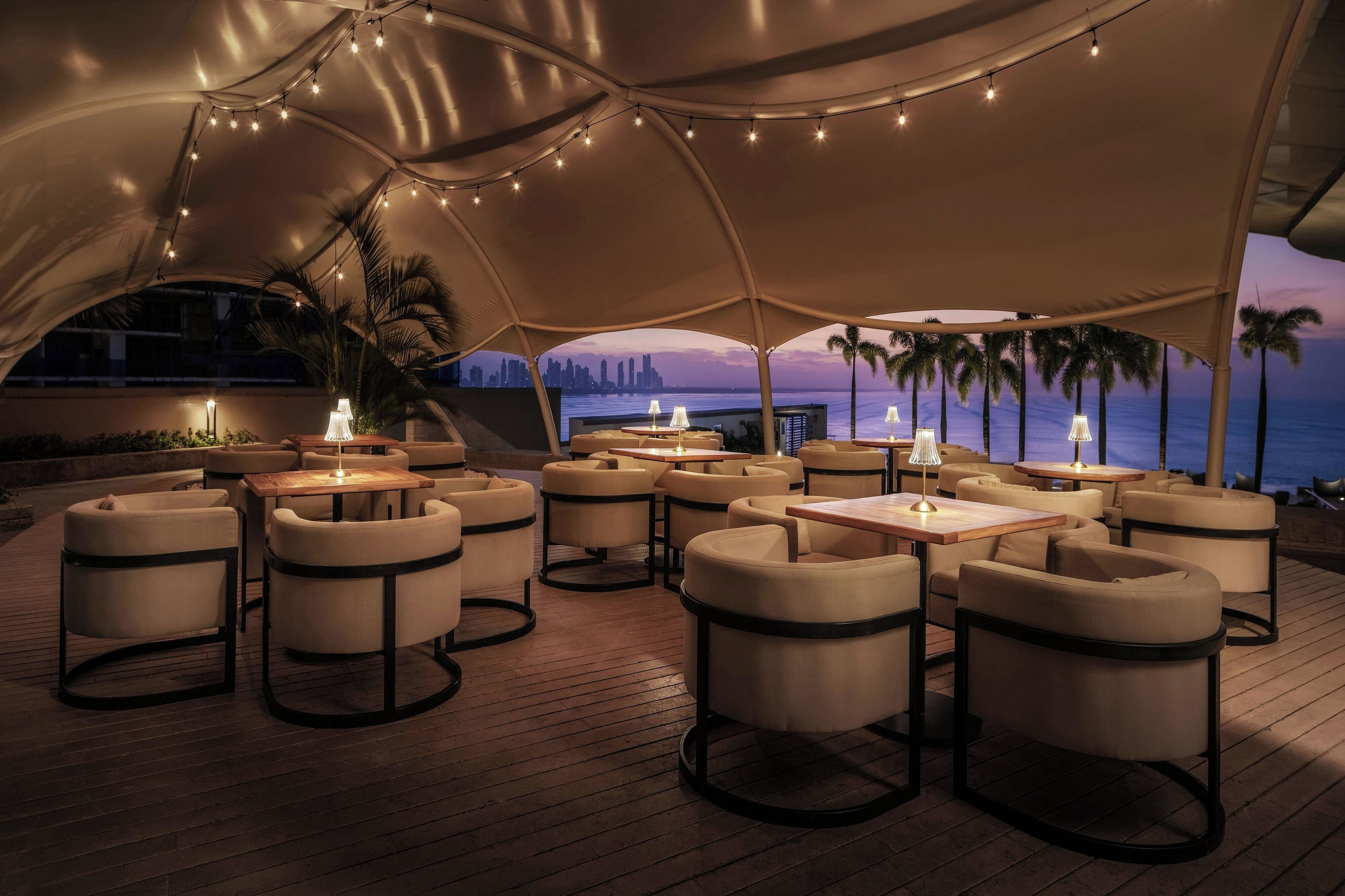 An outdoor seating area with round tables and cushioned chairs, under a large canopy with string lights.