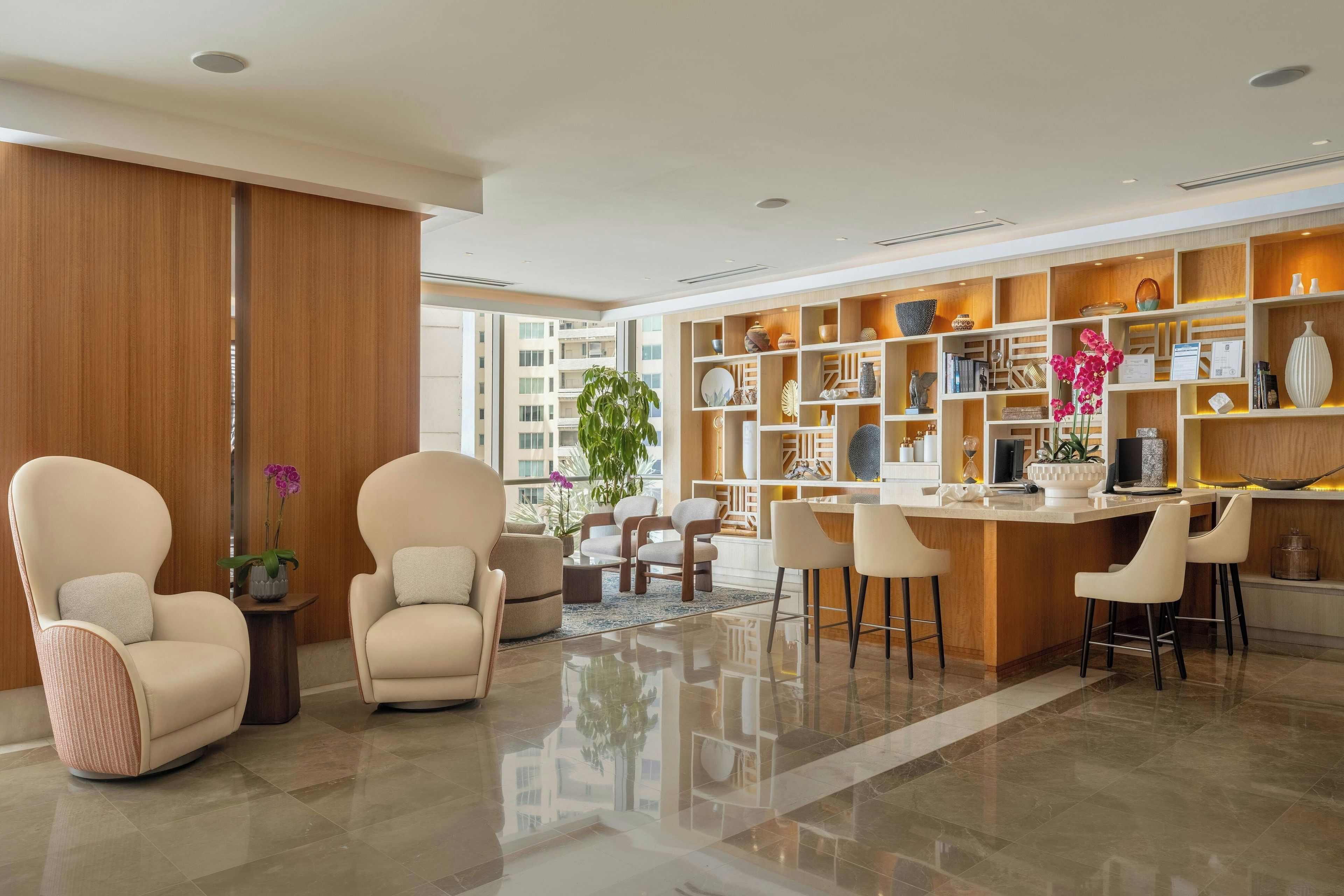 A modern living room with a large window, a wooden bookshelf, and a dining area with bar stools.