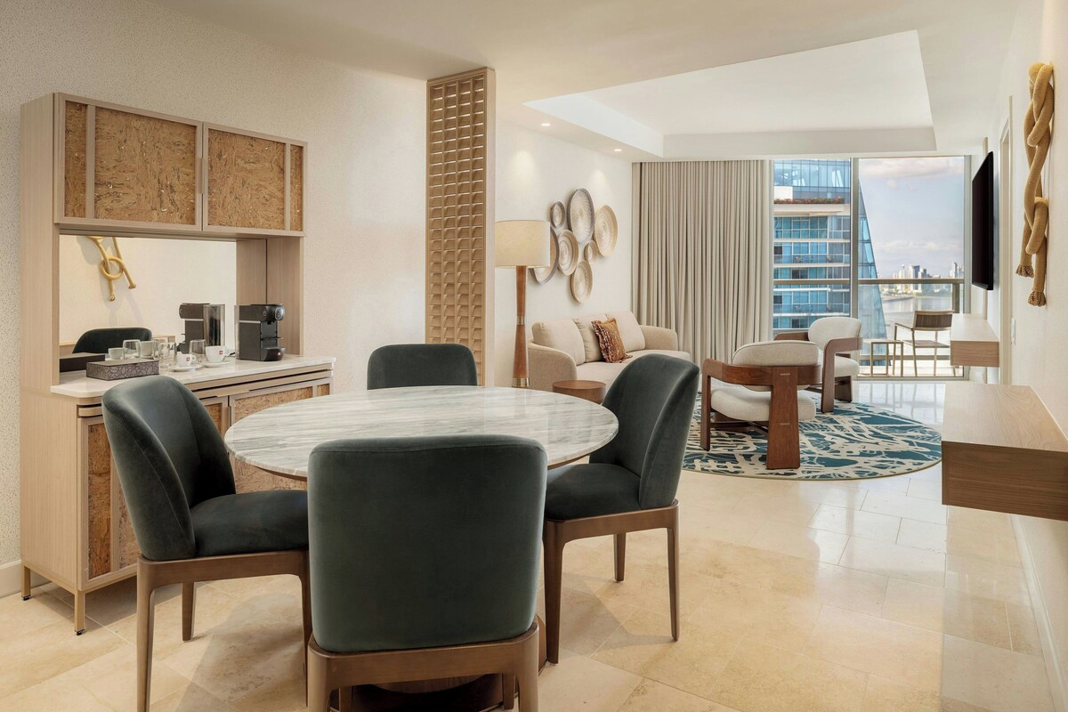 A modern hotel room with a dining area, a sofa set, a TV, and a view of the cityscape.