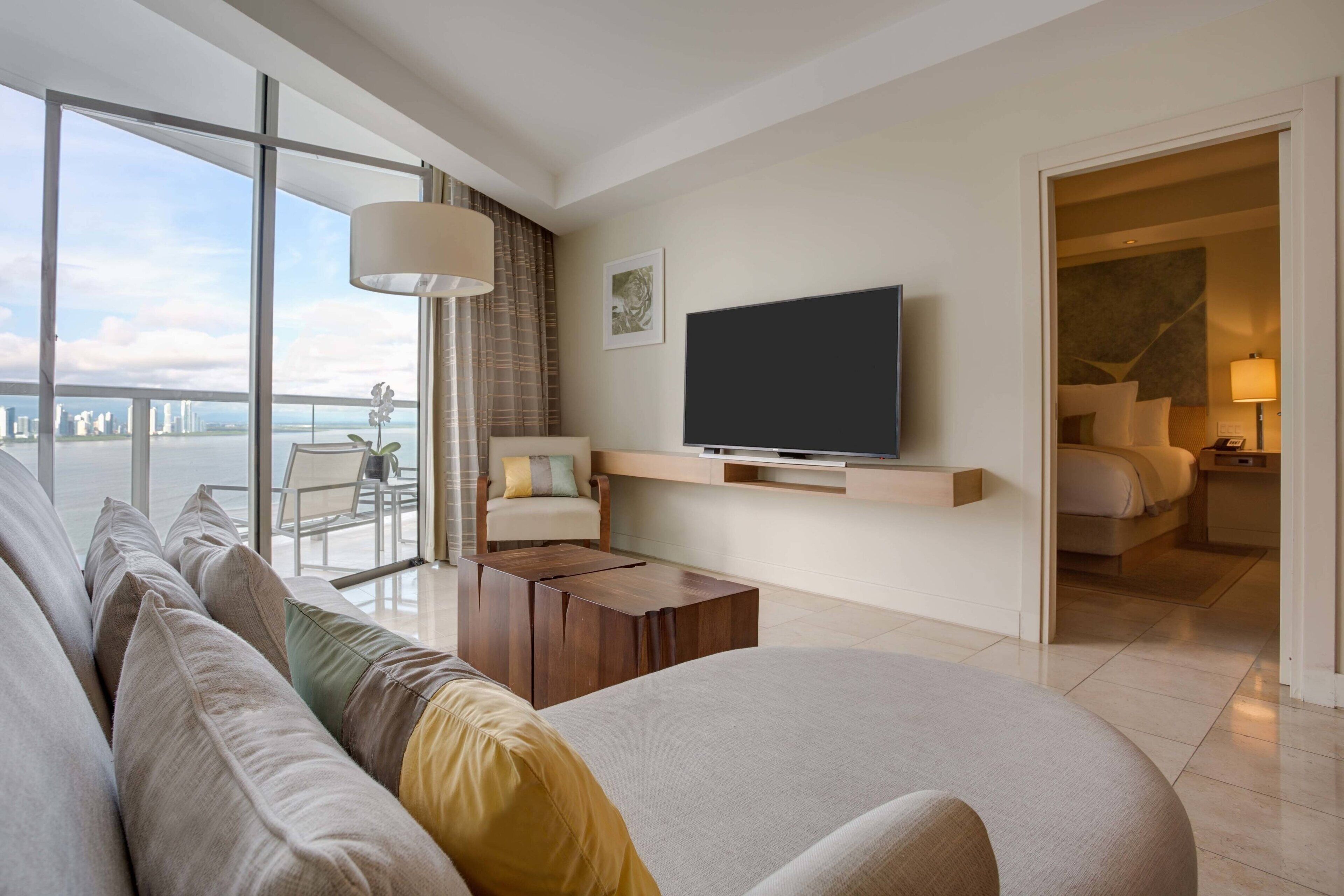 Una sala de estar moderna con un televisor de pantalla plana grande, un sofá, una mesa de centro y un balcón con vistas al horizonte de la ciudad.