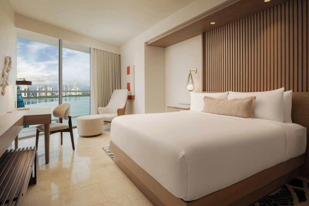 A modern hotel room with a large bed, a desk, a chair, and a view of the cityscape.
