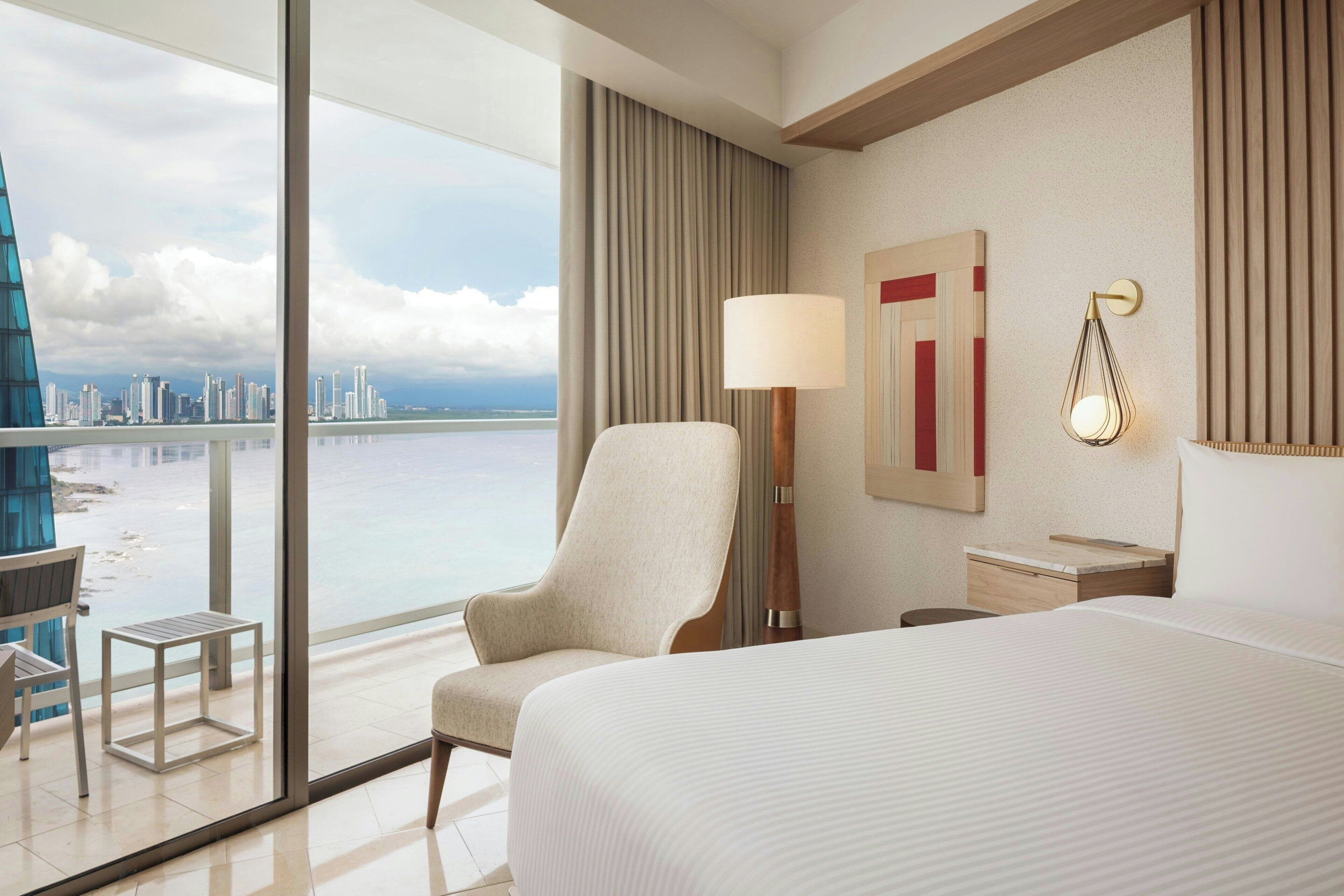 Une chambre d’hôtel avec un grand lit, une chaise, une petite table, une lampe et une vue sur les toits de la ville, visible à travers une grande fenêtre.
