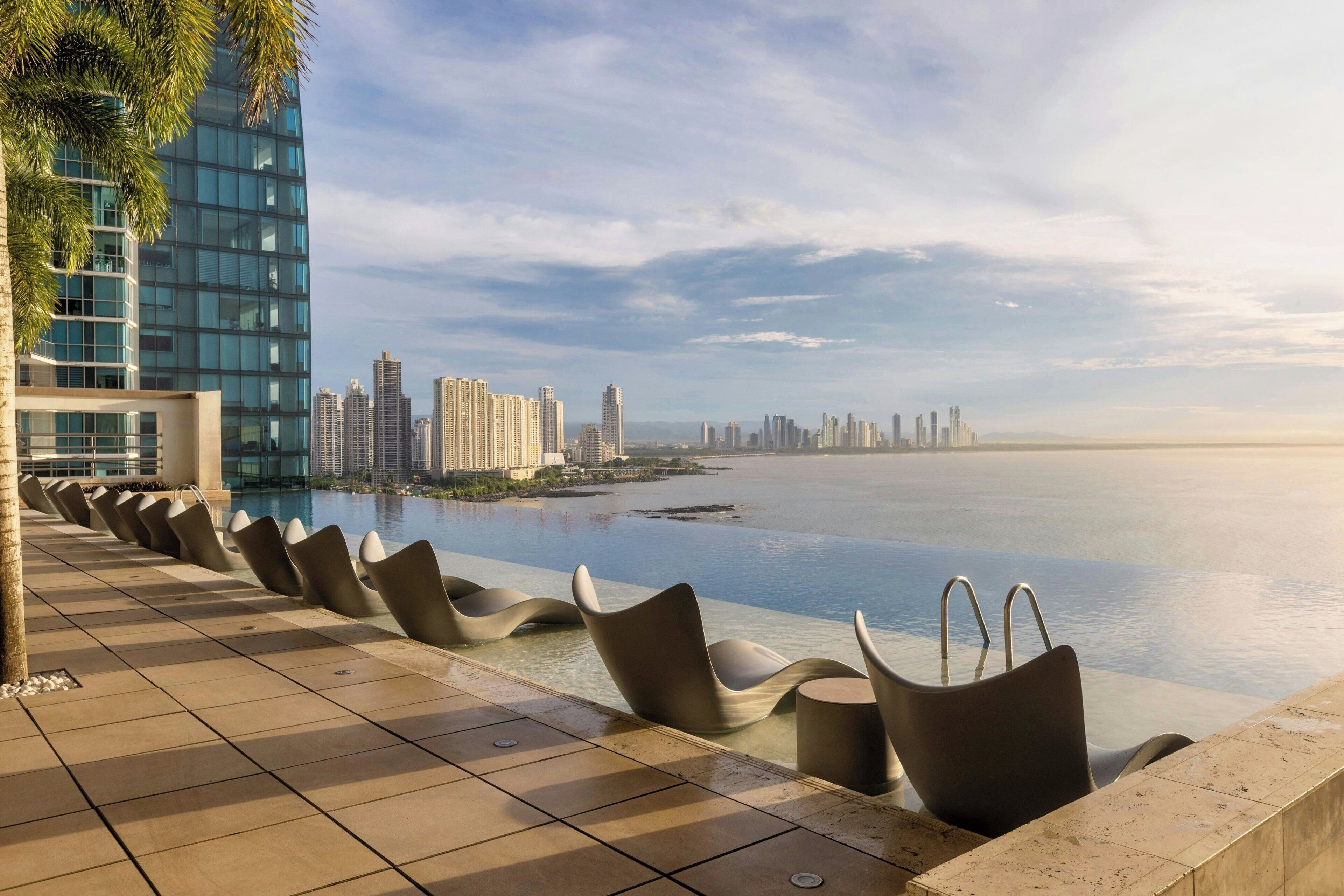 A waterfront terrace with modern chairs, a pool, and a city skyline in the background.