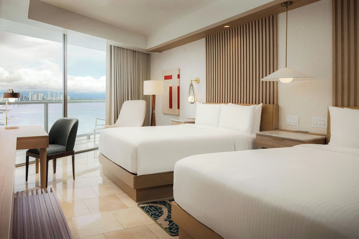 Habitación de hotel con dos camas, un escritorio, una silla, un ventanal grande con vistas a la ciudad y una lámpara moderna.
