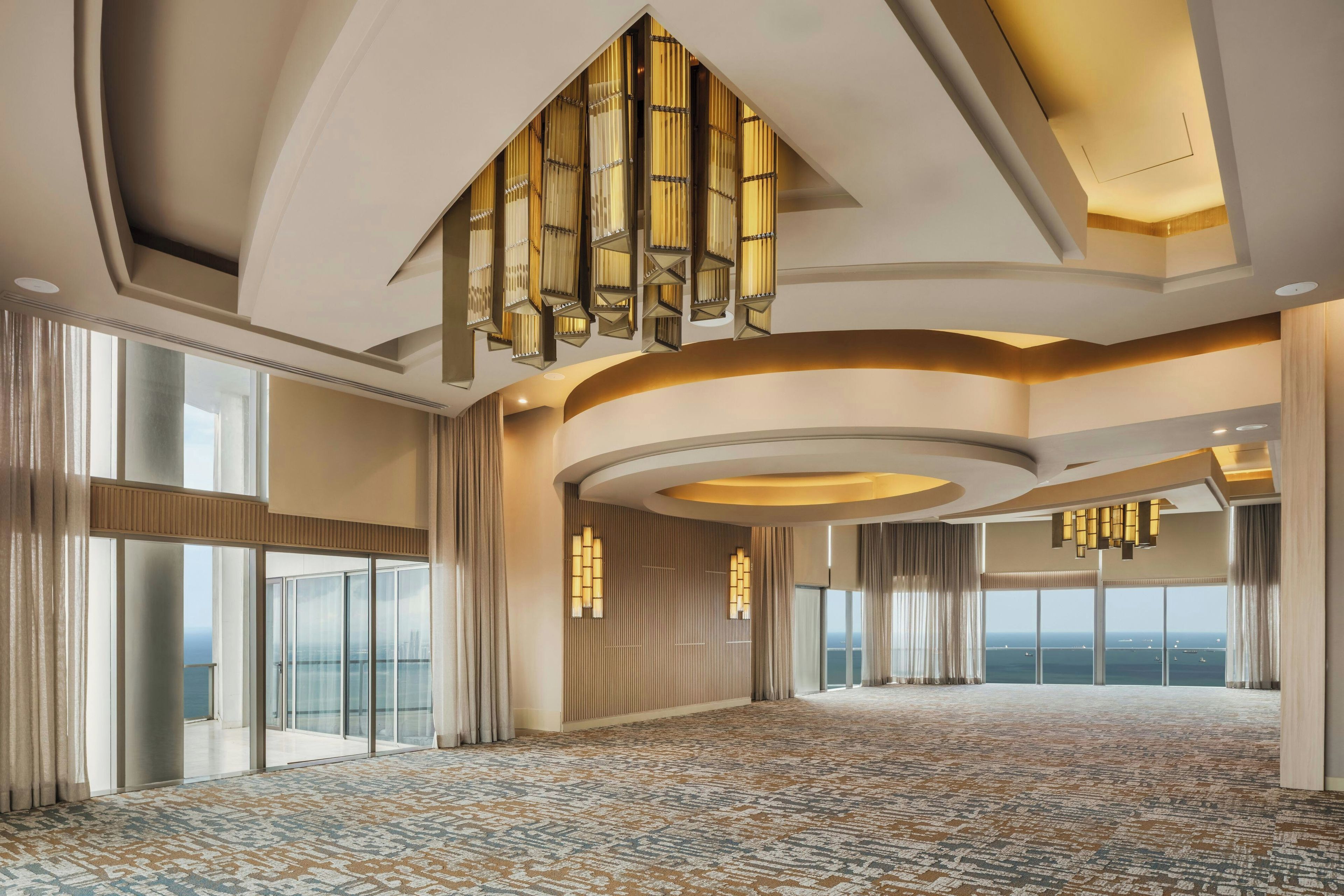 Une chambre d’hôtel spacieuse, dotée de grandes fenêtres, d’un tapis à motifs et d’un lustre moderne.