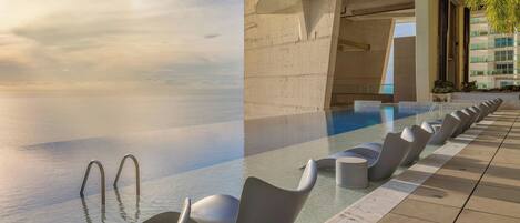 Una piscina infinita con sillas modernas y una estructura de hormigón al fondo.