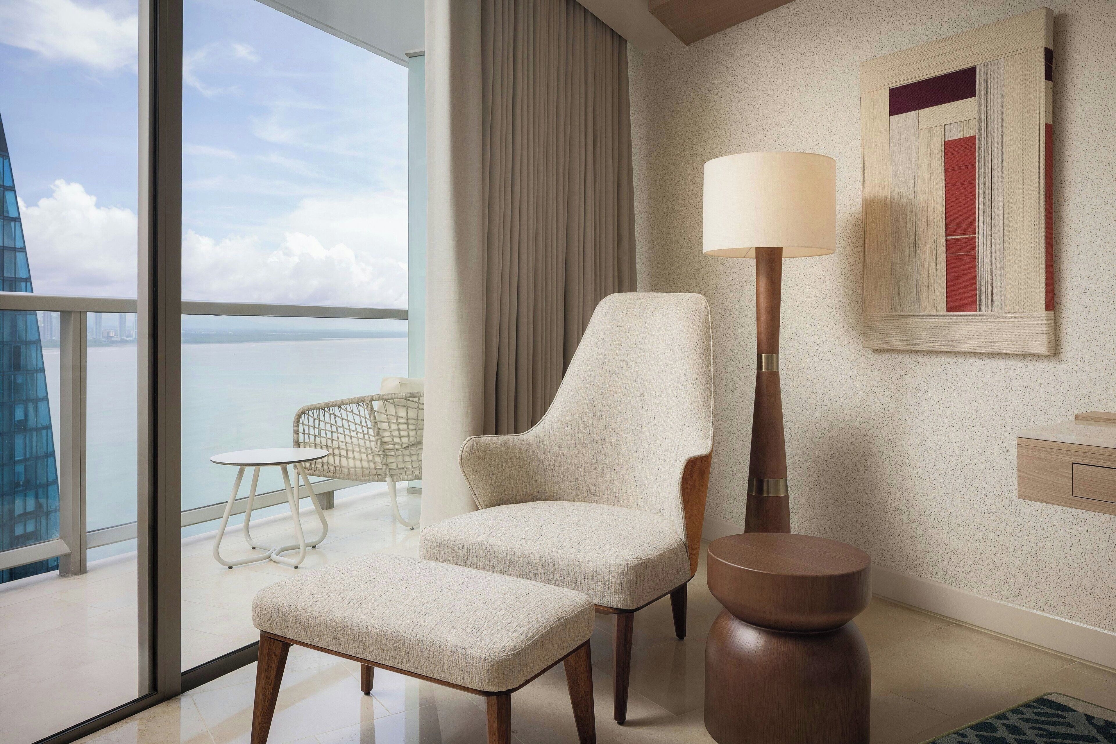 Quarto de hotel moderno com uma grande janela, uma poltrona confortável, uma mesinha, uma luminária e uma obra de arte emoldurada na parede.