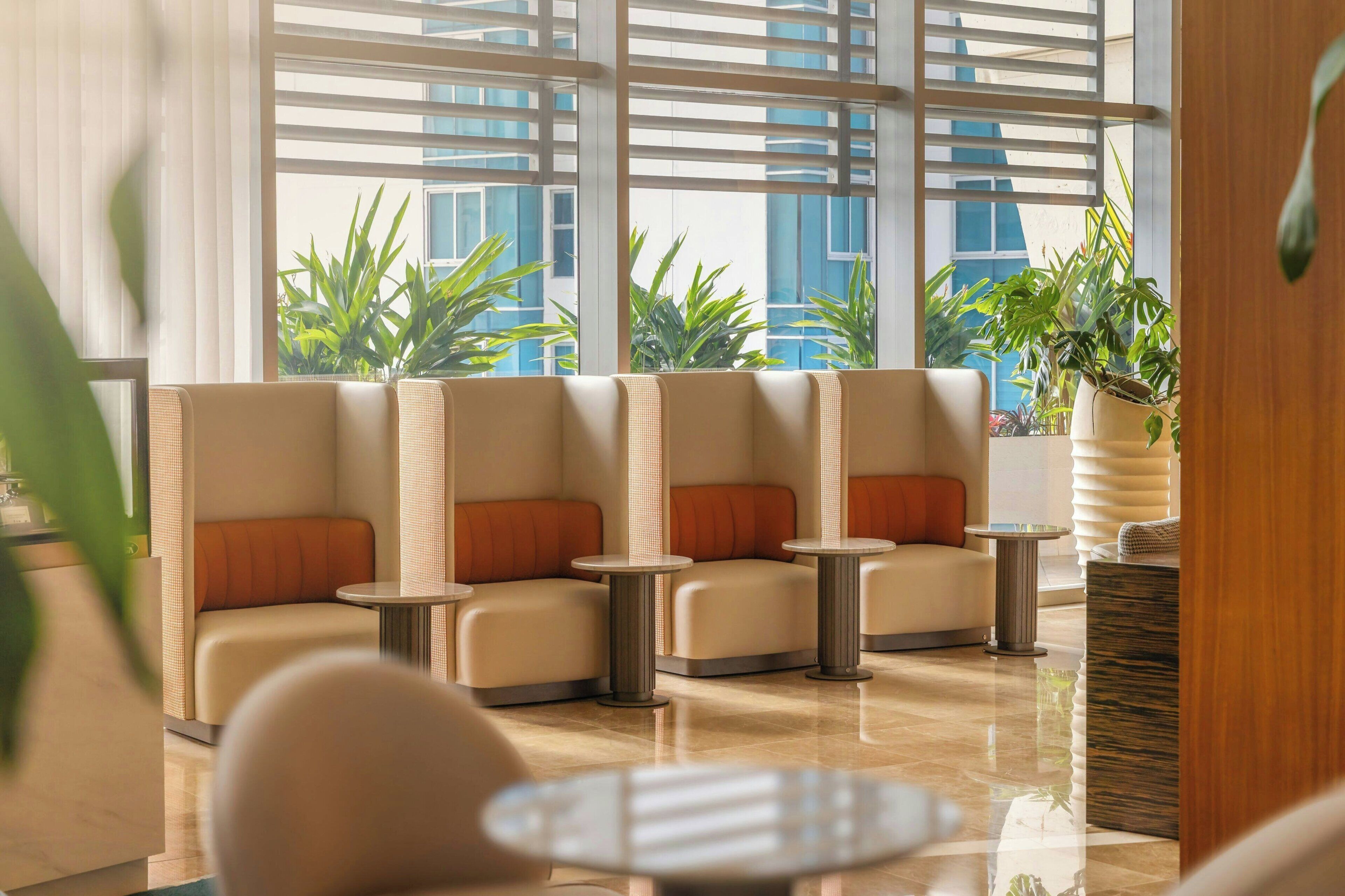 Un área de estar con sillones acolchados y mesitas pequeñas, ventanas amplias con persianas y plantas de interior.