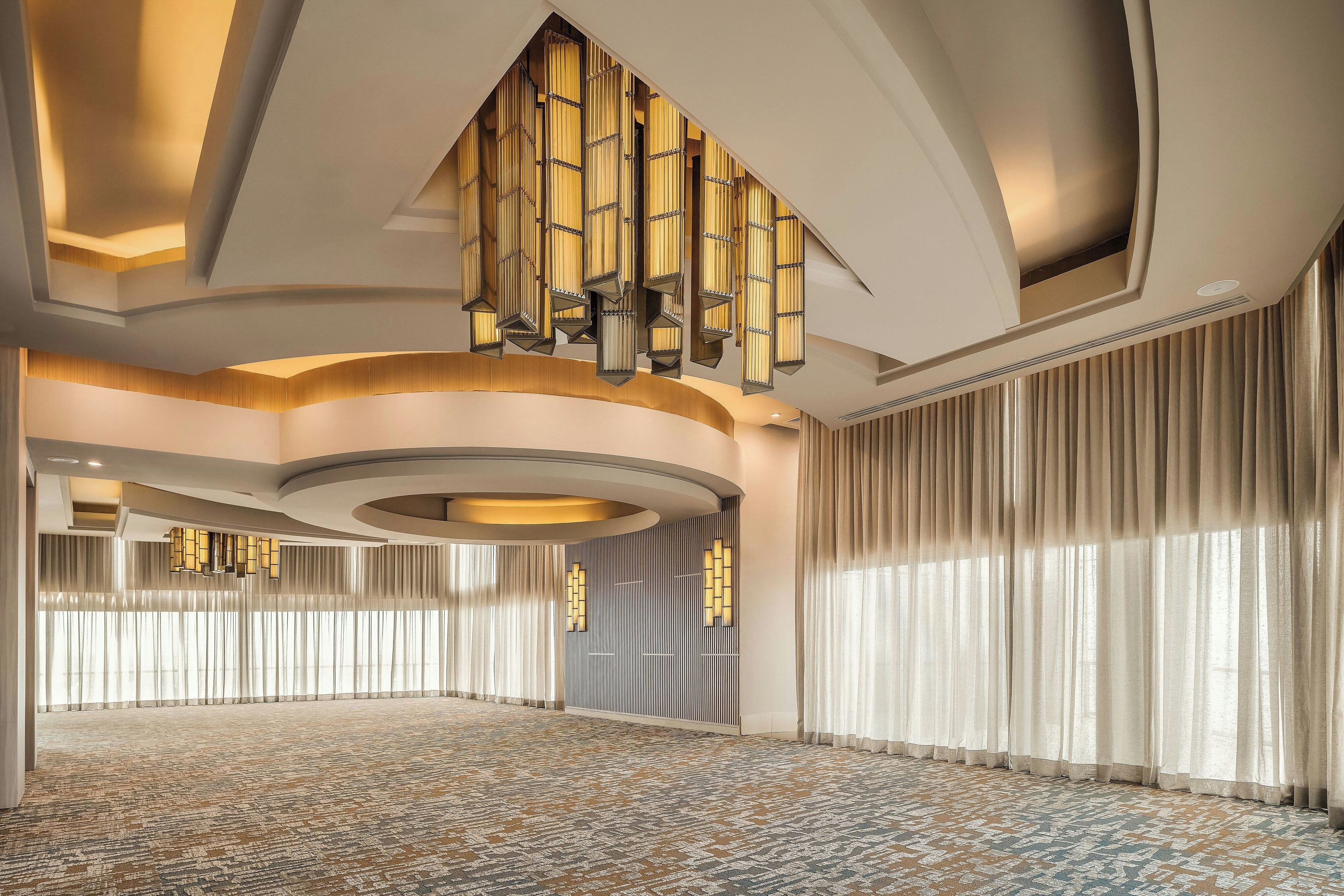 Amplia habitación de hotel con alfombra estampada, ventanas grandes con cortinas y un diseño de techo original con iluminación empotrada.