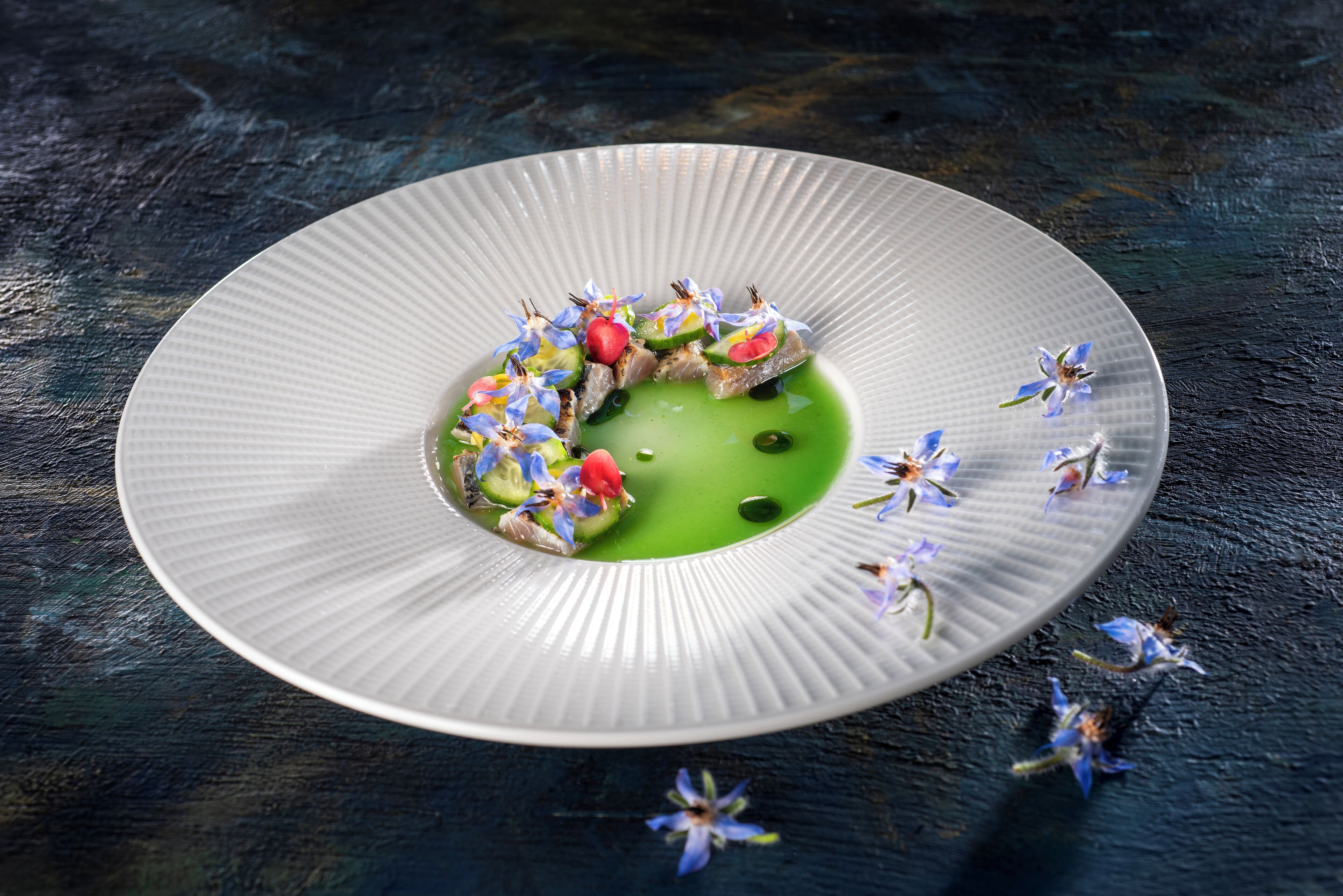 Un piatto bianco con salsa verde e diversi cibi, tra cui un frutto rosso e fiori blu, su una superficie scura e strutturata.