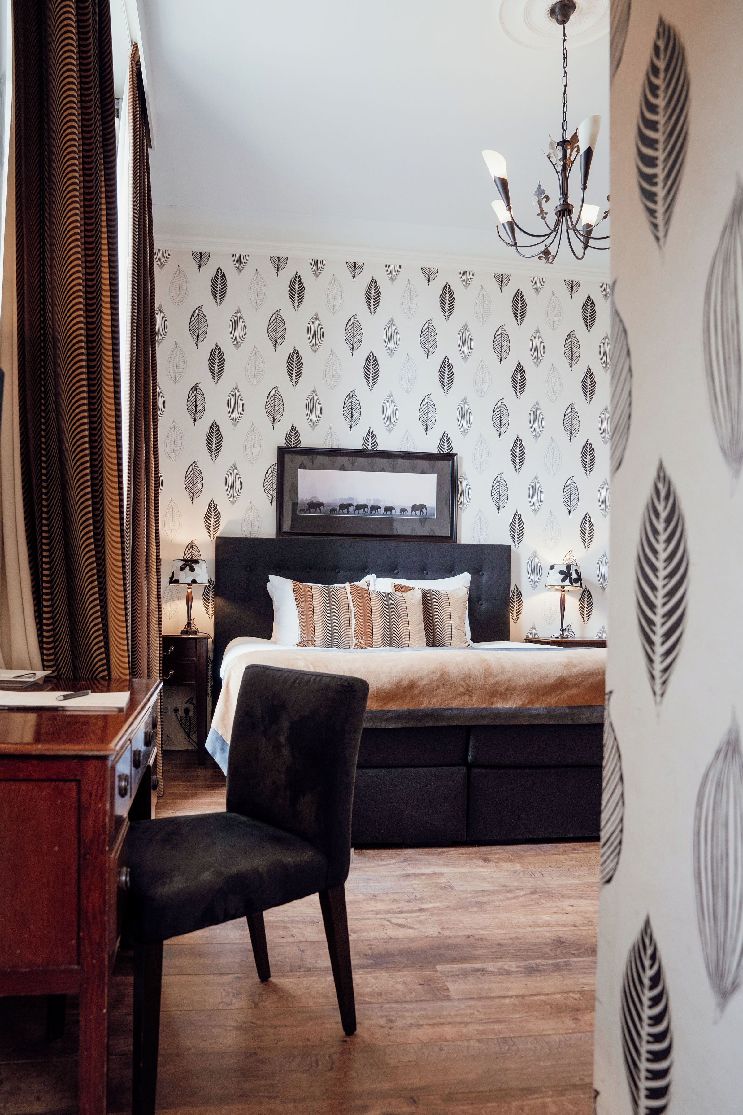 A bedroom with a bed, a desk, a chair, a chandelier, and leaf-patterned wallpaper.
