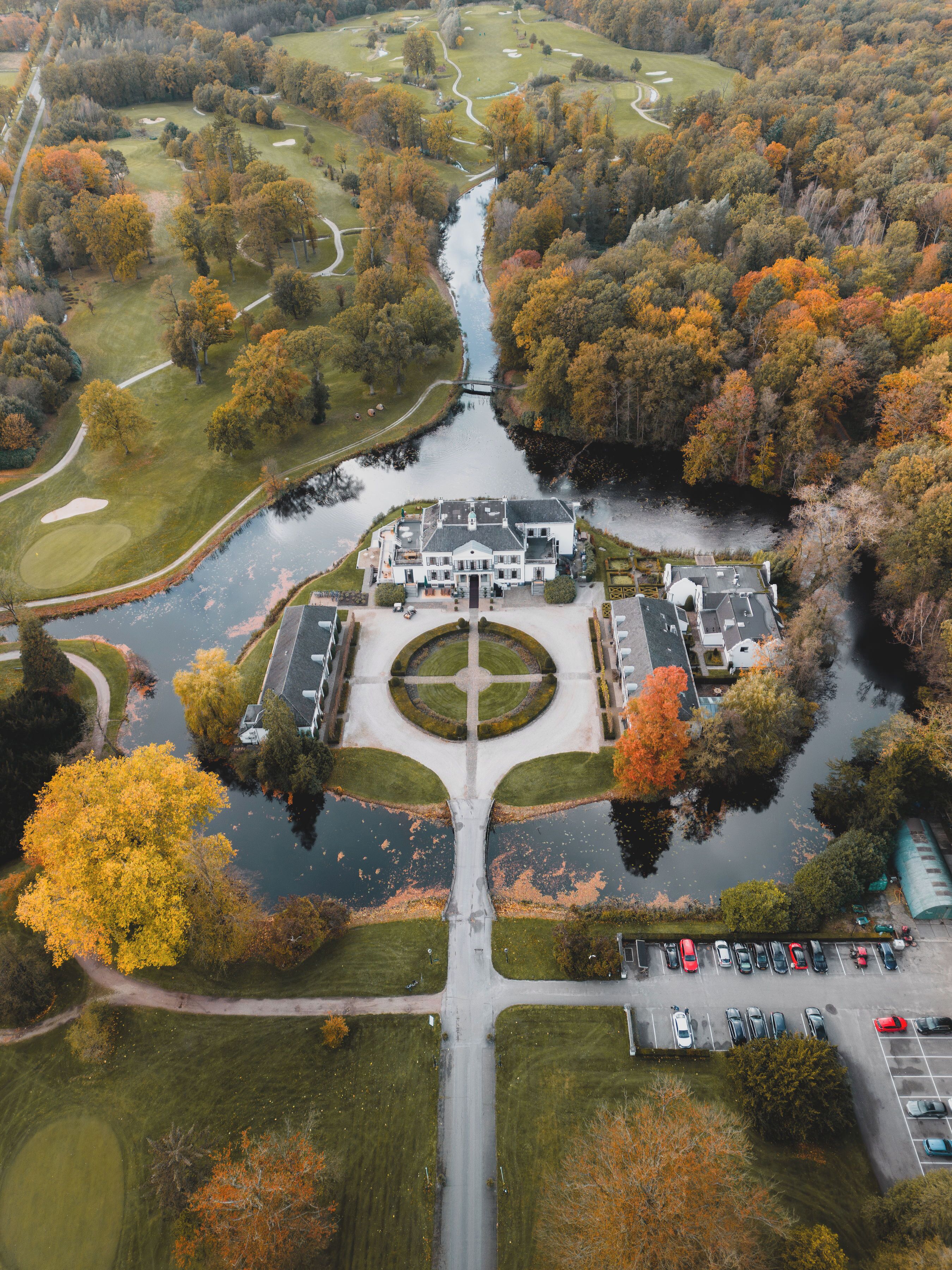 An aerial view of a golf course with a clubhouse, a central water feature, and parking areas.