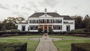 Un edificio grande y simétrico con una entrada central, flanqueado por jardines cuidados y un camino de adoquines que conduce a la entrada.