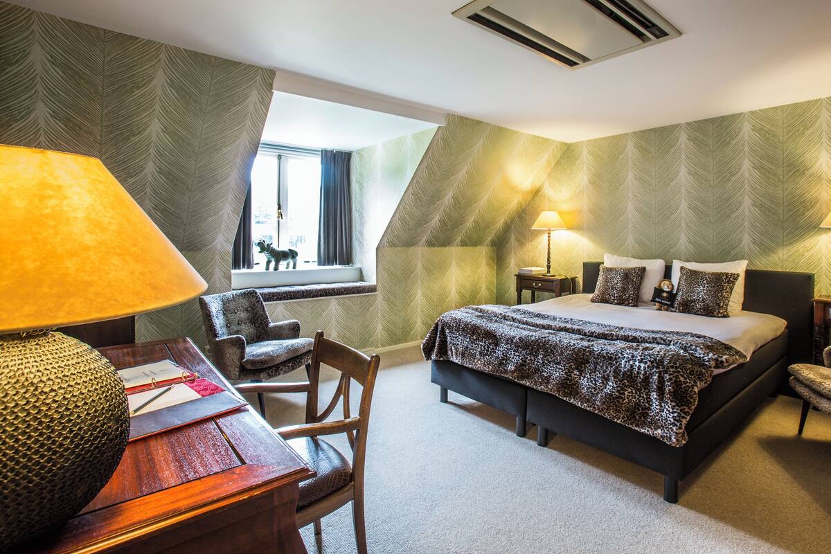 Habitación de hotel con cama, dos mesitas de noche, un escritorio con lámpara, una silla y ventana con vista a un perro afuera.