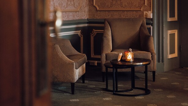 A room with a patterned wallpaper, a brown armchair, a smaller chair, and a small table with a candle. - Kasteel Engelenburg - Quality Lodgings (Brummen)