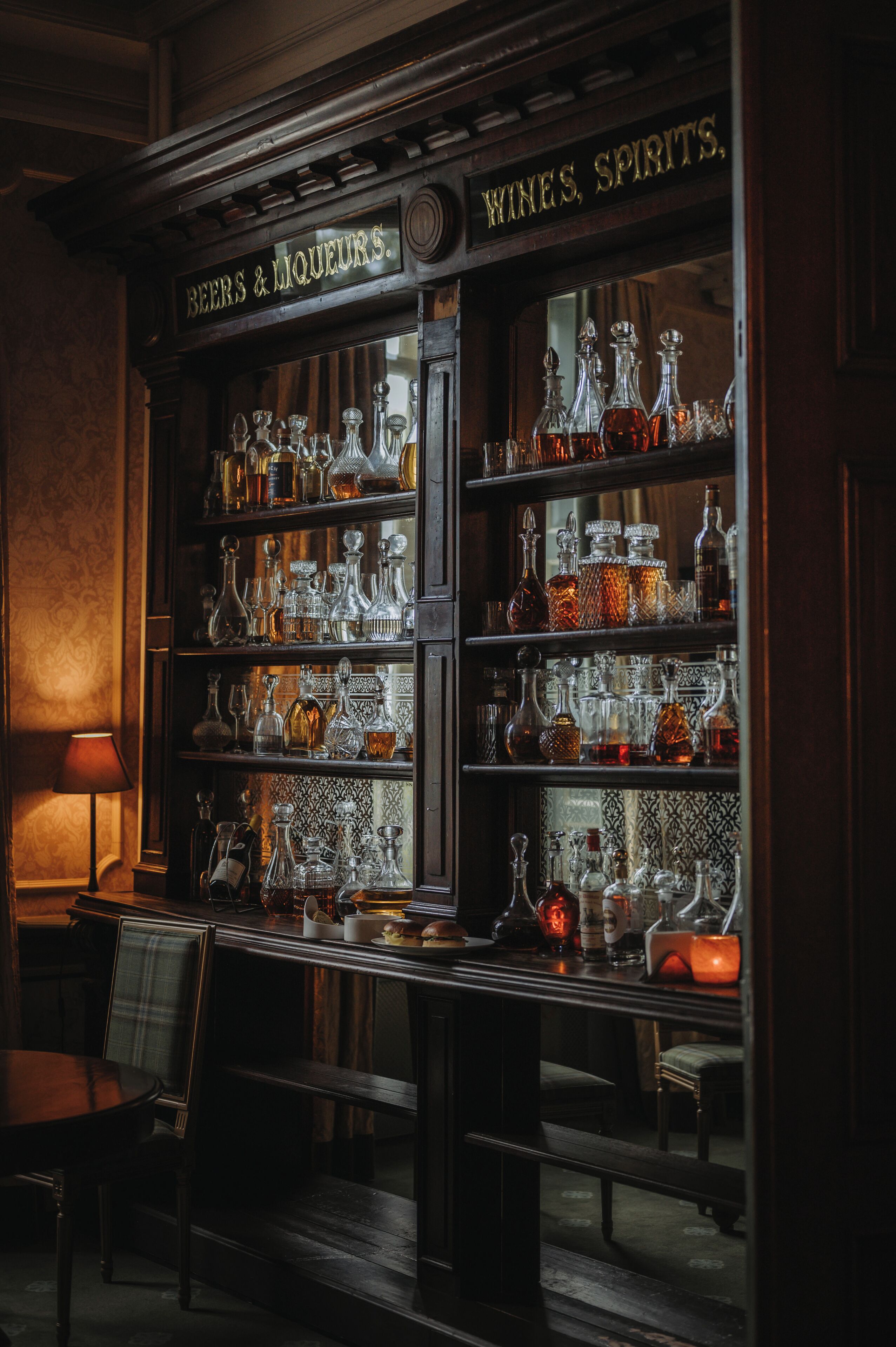 Un espace bar avec des étagères remplies de bouteilles et de verres de toutes sortes, une enseigne indiquant « Bières et spiritueux, vins », et un banc avec des chaises.